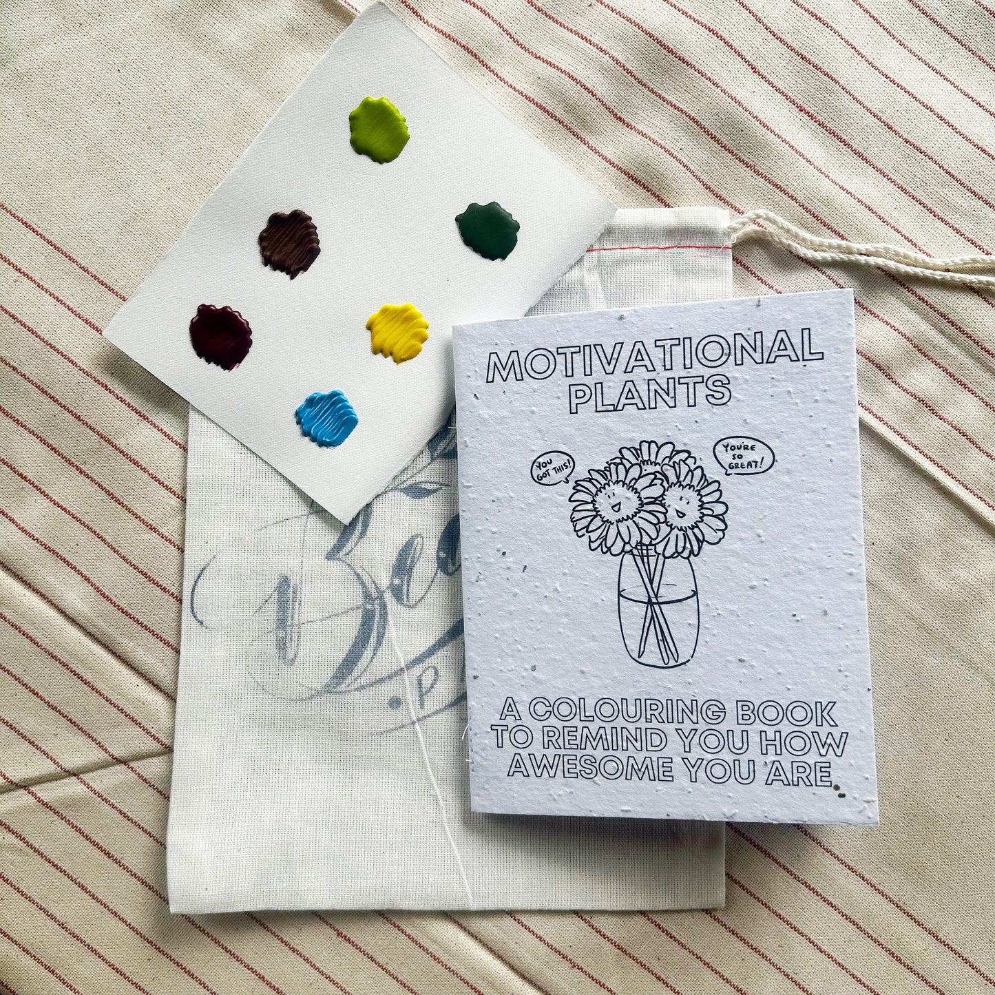 A coloring book titled 'Motivational Plants' with a palette of six watercolor colors and a brush, displayed on a striped background.