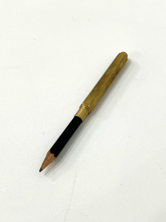 A short black wooden pencil inserted into a cylindrical brass pencil extender, placed on a plain white surface. The extender has a smooth metallic finish and adds length and grip to the pencil stub.