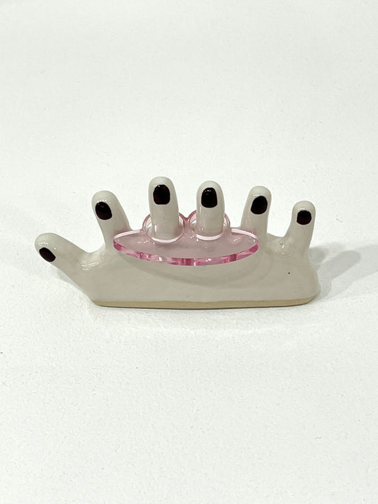 A ring with a hand-like design, featuring a pink central element and black details on a white background.