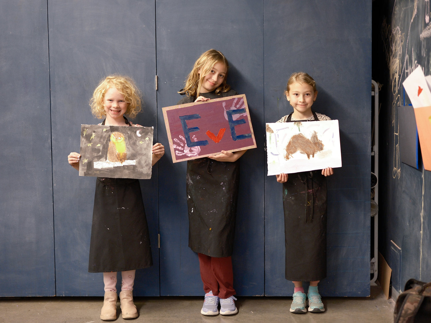 Three young girls standing in front of a blue wall, each holding up a piece of artwork they have created, with various colors and textures visible on their pieces.